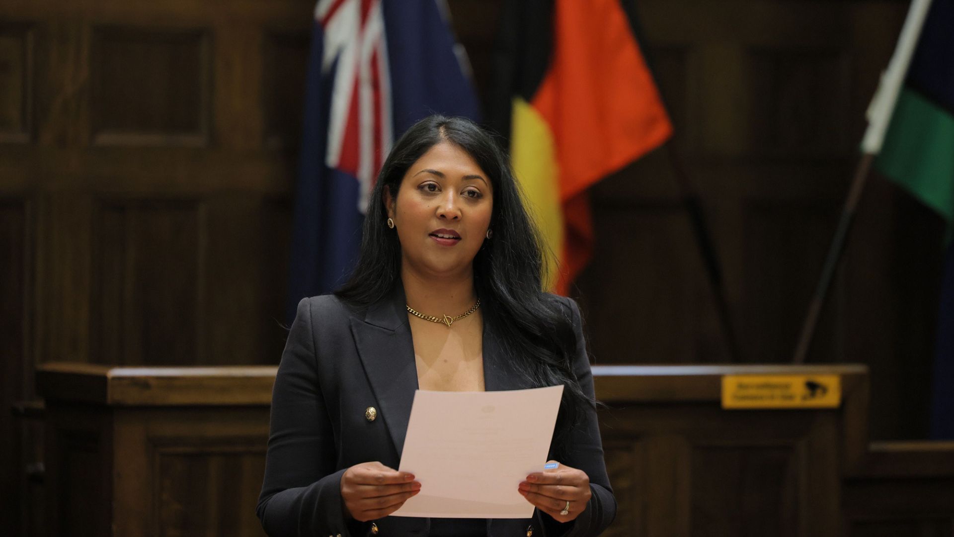 Sam Choudhury giving speech in council chamber, Australian and Aboriginal flags behind her