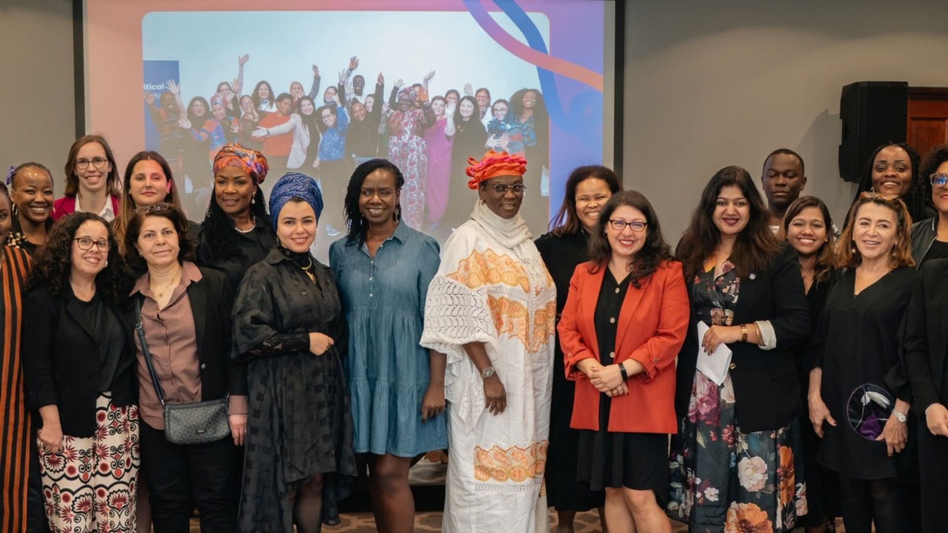 Non-professional photo pf a group of diverse women smiling and posing. Some are wearing African headdress, others in suits and dresses