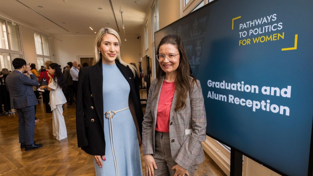 Two smartly dressed women stand next to a digital sign reading "Graduation and Alum Reception"