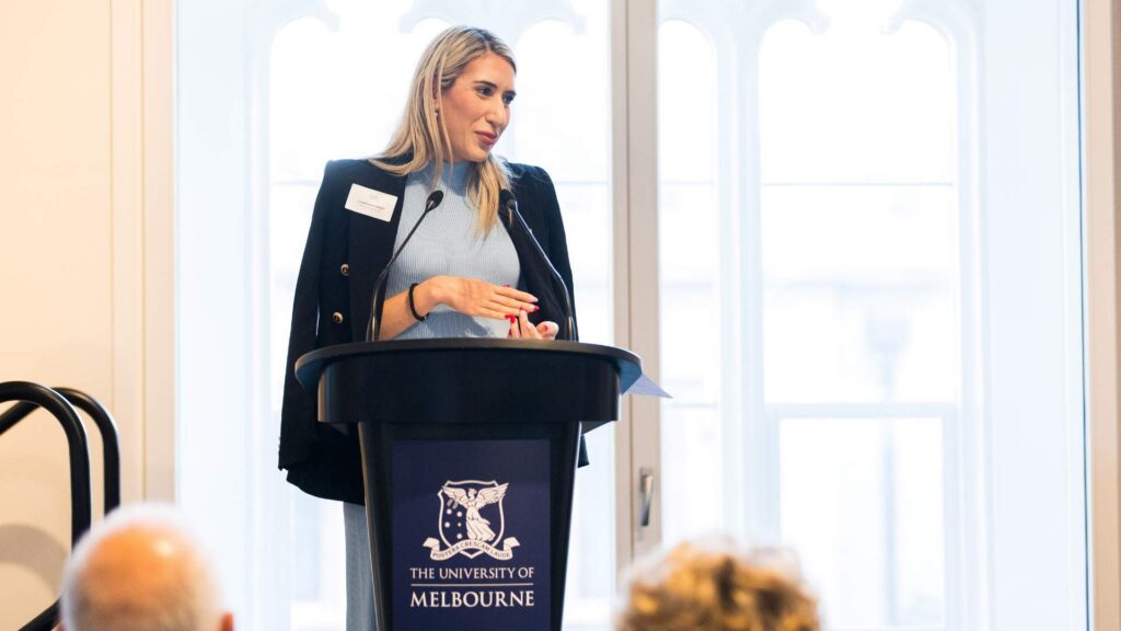 Candid photo of Katharine Nikolic, a young blonde woman speaking at a podium with the University of Melbourne logo. She has a navy blazer over her shoulders