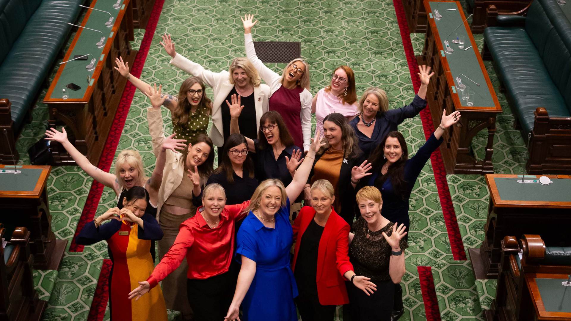 Large group of women grinning with their arms spread wide, inside the South Australian parliament chamber