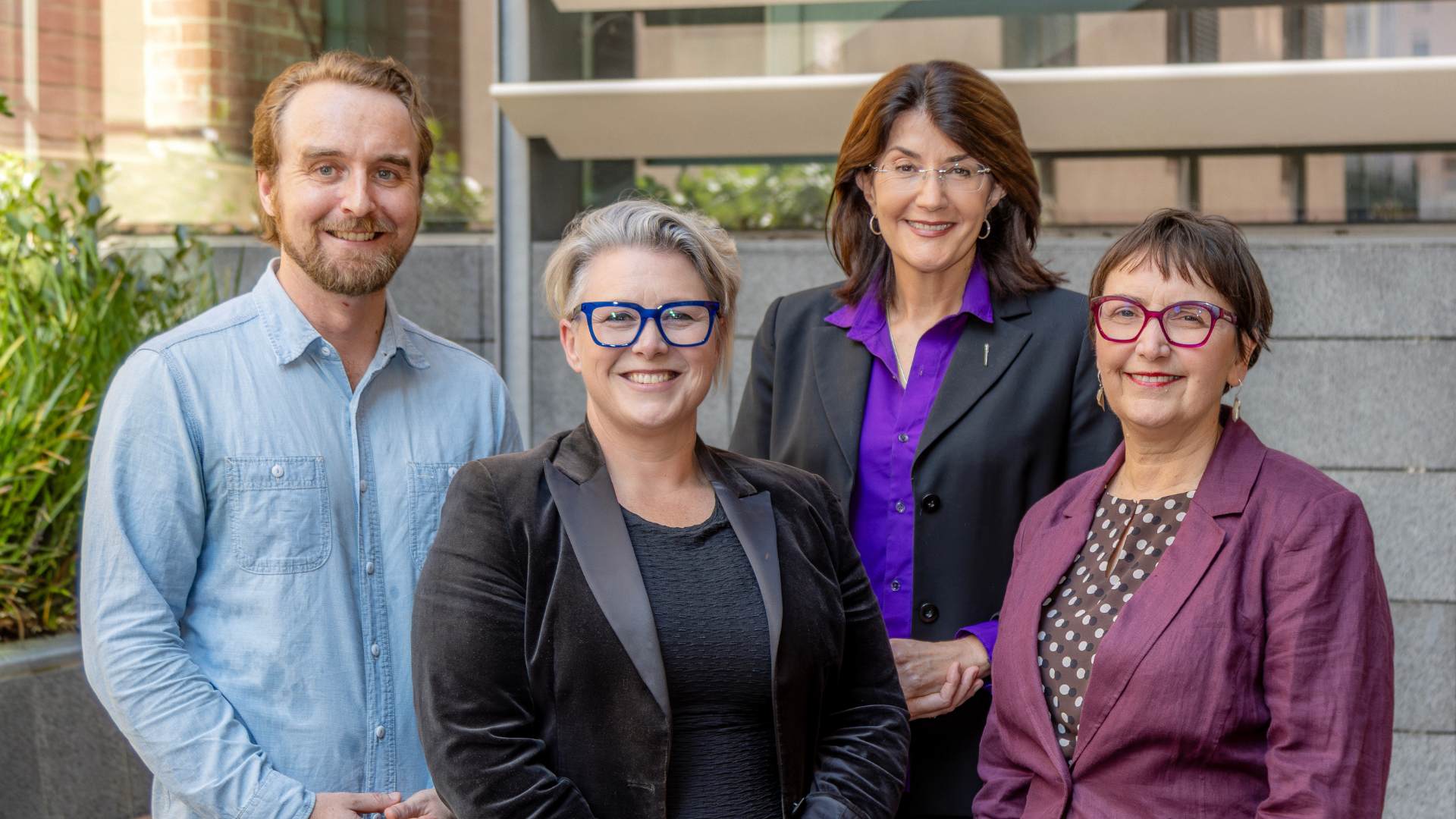 Three women and one man, dressed in shirts and blazers, smile outside a public building