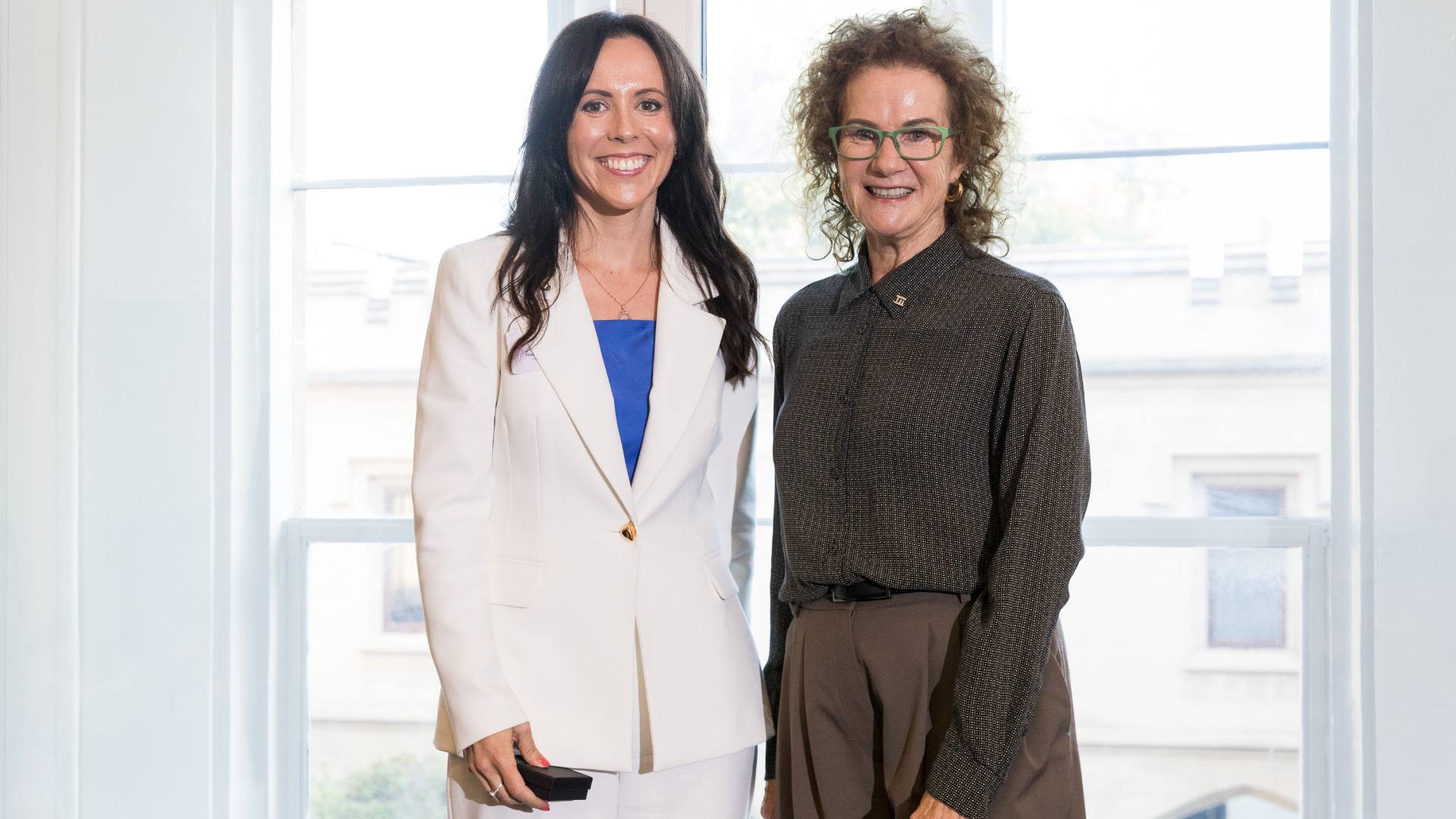 Photo of two women smiling and posing together: one has long dark hair and is wearing white suit, the other has shorter curly hair and glasses and is wearing a brown shirt and pants