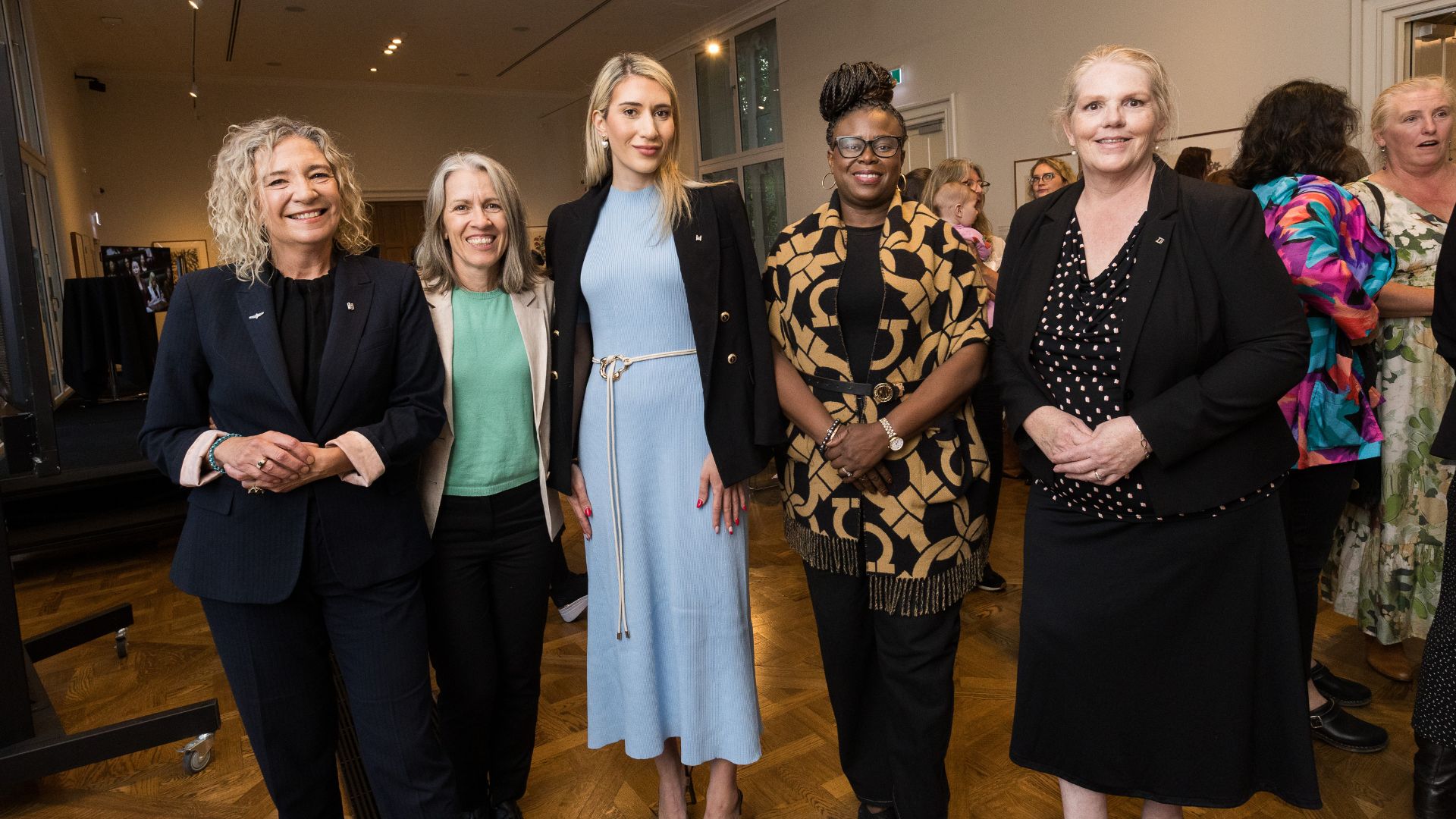 Group of 5 diverse, well dressed women pose for a photo in a large room