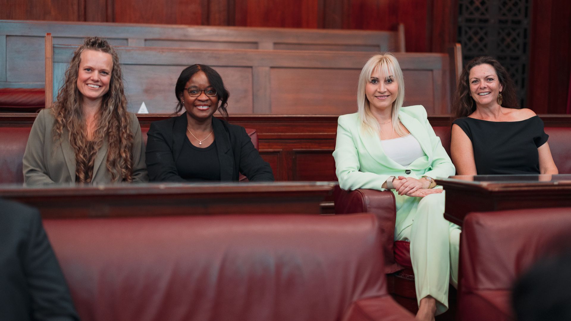 Photo of four diverse smiling women sitting in wood and leather bench seats in parliament