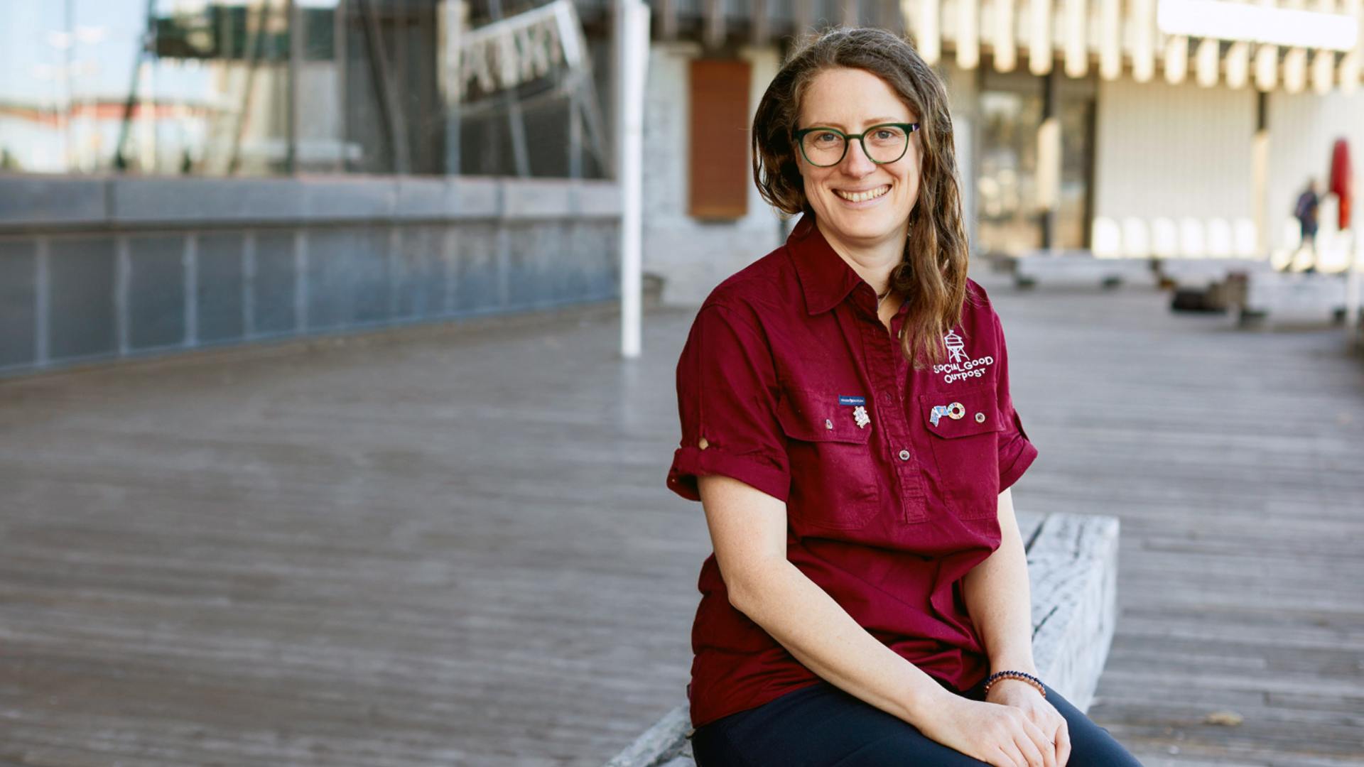 Photo of Lara Stephenson wearing glasses and a burgundy short sleeved shirt, siting outside a modern glass and concrete building