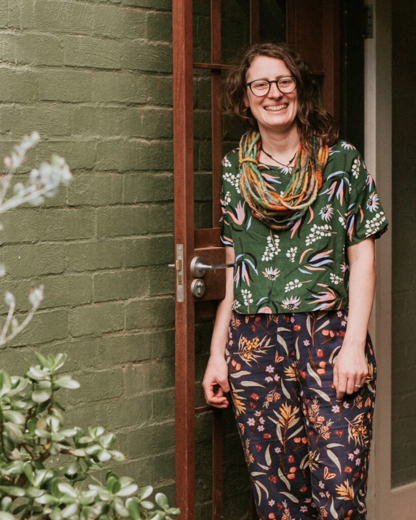 Photo of Lara Stephenson wearing glasses, short sleeved shirt and pants with native plant print, standing outside a green brick house