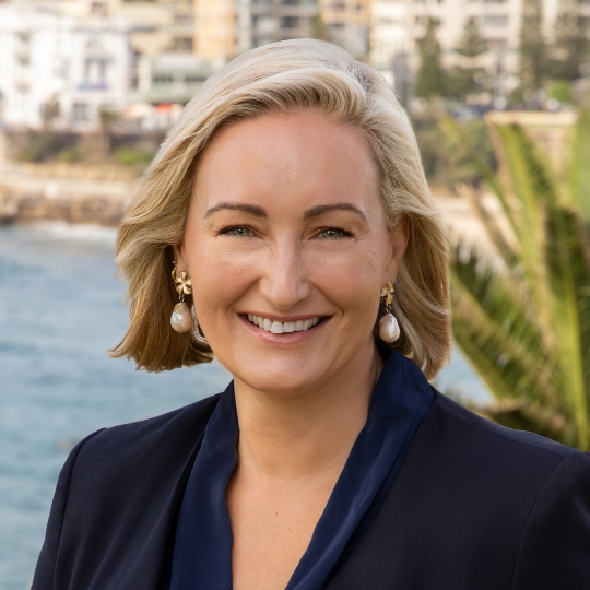 Headshot of Dr Marjorie O'Neill outdoors, sea and town in the background, wearing a black blazer