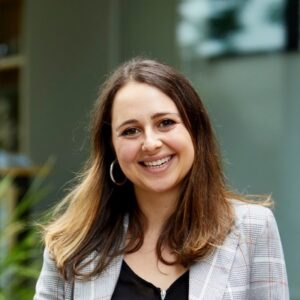 Photo of Ashleigh Streeter-Jones, a smiling young woman with long brown hair, wearing a gray blazer and hoop earrings