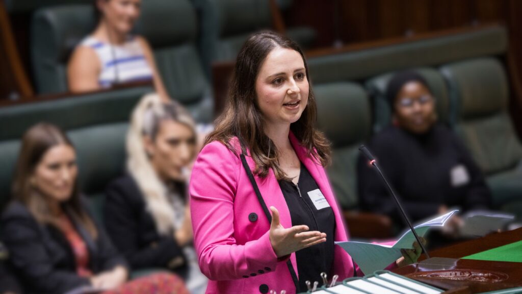 Photo of Asheligh Streeter-Jones, a young white woman with long brown hair, wearing a bright pink blazer. She is standing in the Victorian parliament giving a speech in the centre while women behind her watch