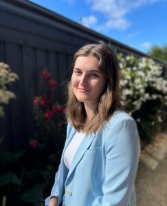 photo of Ellie Mueller, a young white woman with long light brown hair. She is outside a house with floral shrubbery and a blue sky behind her.