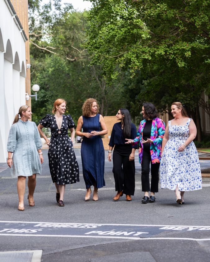 Six smartly dressed women walking and chatting, trees in the background