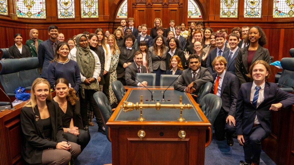 group of diverse teenagers posing in rows inside the WA parliament, surrounded by ornate wood panelling and stained glass