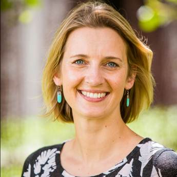 Photo of Gabrielle Appleby, a caucasian woman with bobbed blonde hair wearing a black and white blouse and green earrings. There is out of focus greenery in the background.