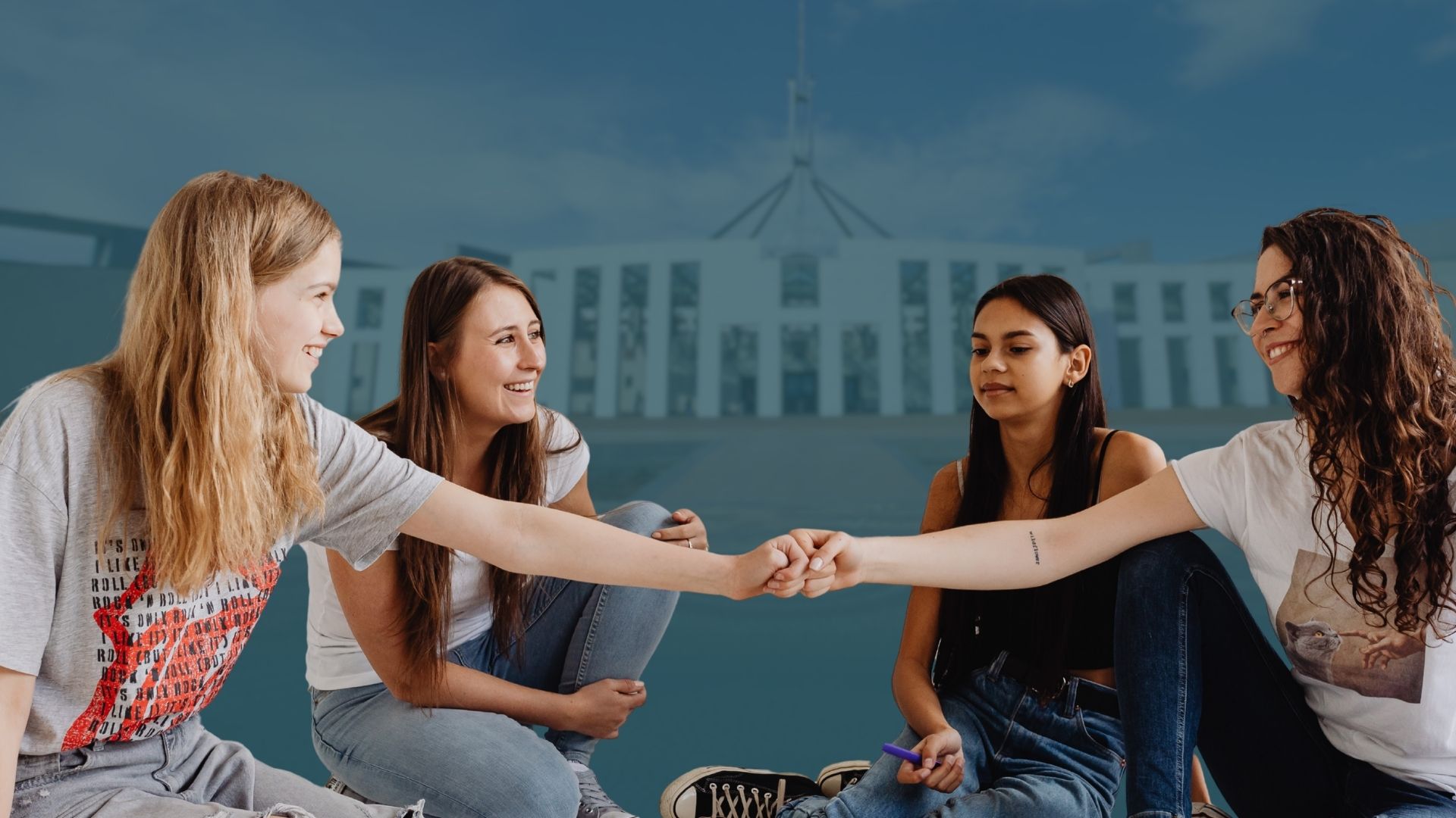 photo of four teenage girls, parliament house of australia in the background. the girls are smiling, dressed casually. Two girls in the centre are bumping fists.