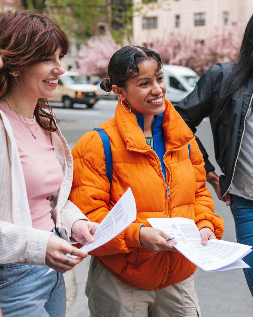 two young women volunteering outdoors, holding papers and smiling