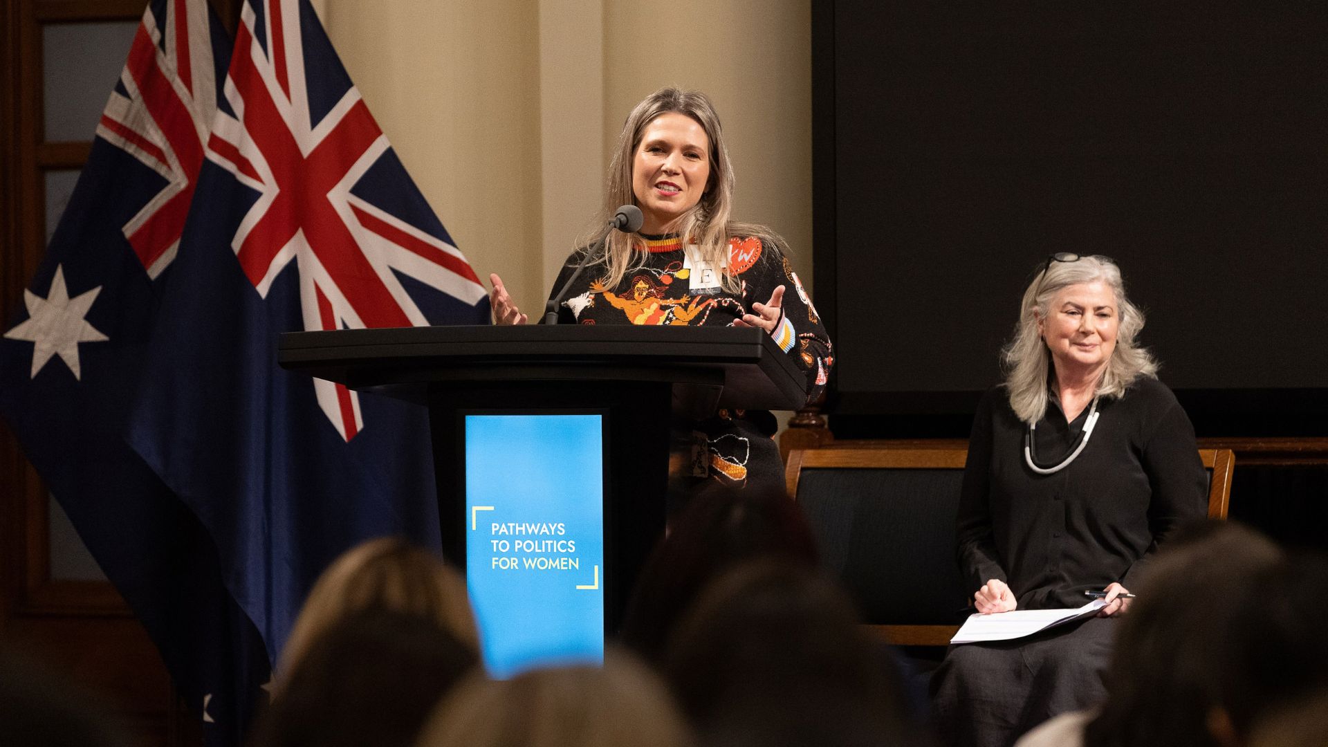 Photo of Jenna Davey-Burns speaking at a lectern with the Pathways to Politics logo on it. She has long blonde hair and is wearing a knit jumper with modern Aboriginal designs on it. There is a large australian flag in the background.
