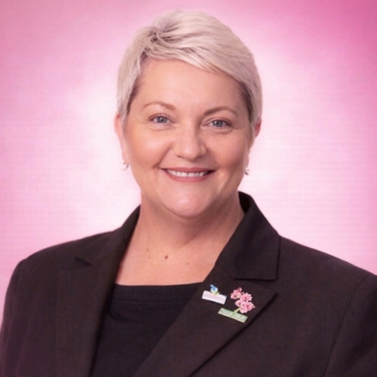 Smiling photo f Leanne aganst a pink background. She has short blonde hair and is wearing a black blazer.