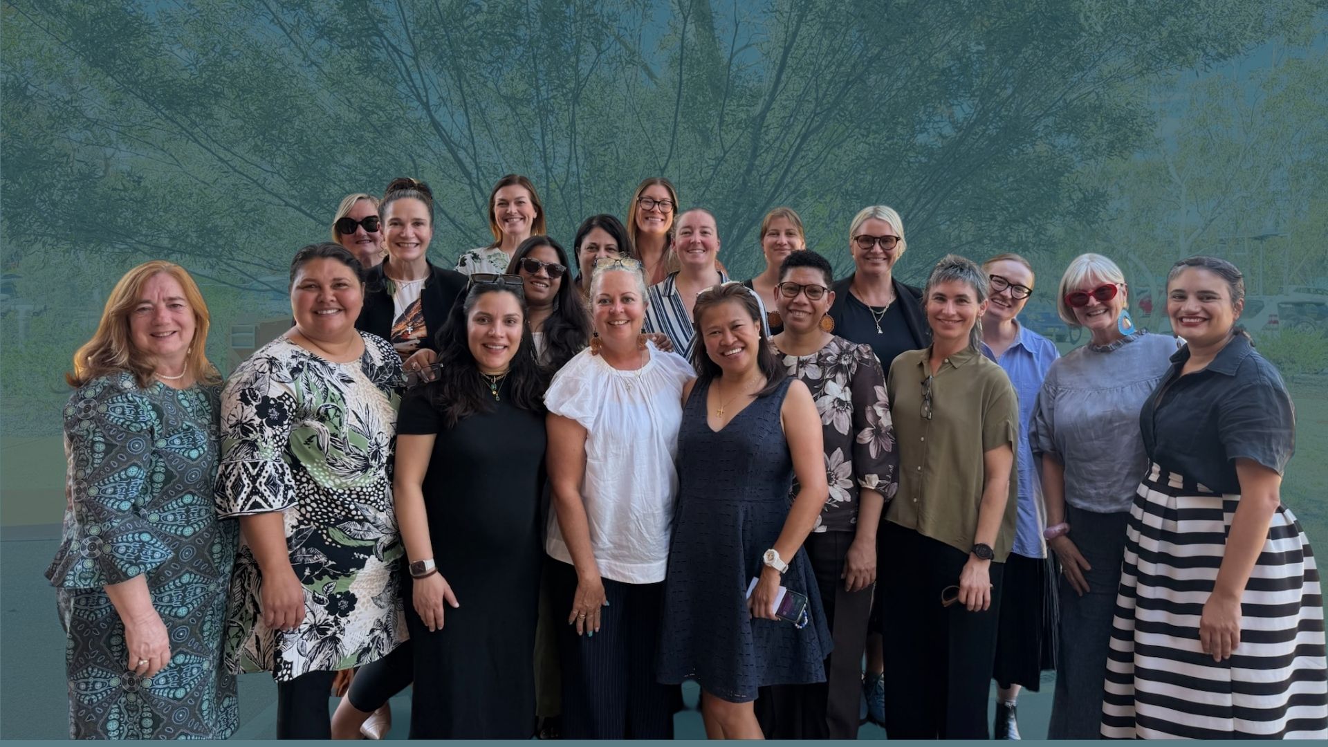 Diverse group of women posing for a photo outdoors, wearing summery clothing, trees behind them