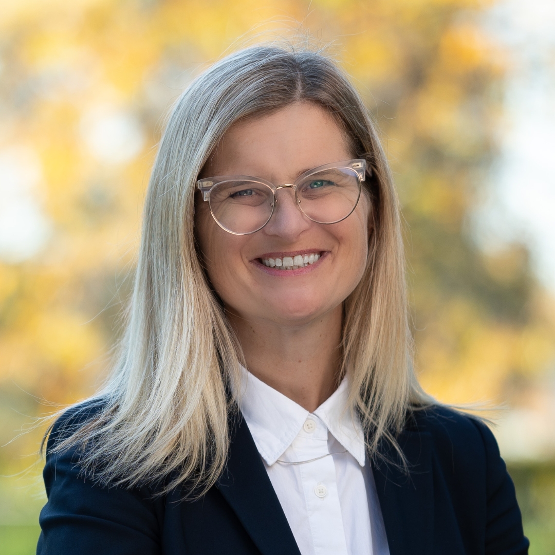Smiling photo of Susanne Newton outdoors, autumnal trees in the background. She has long blonde hair and glasses.