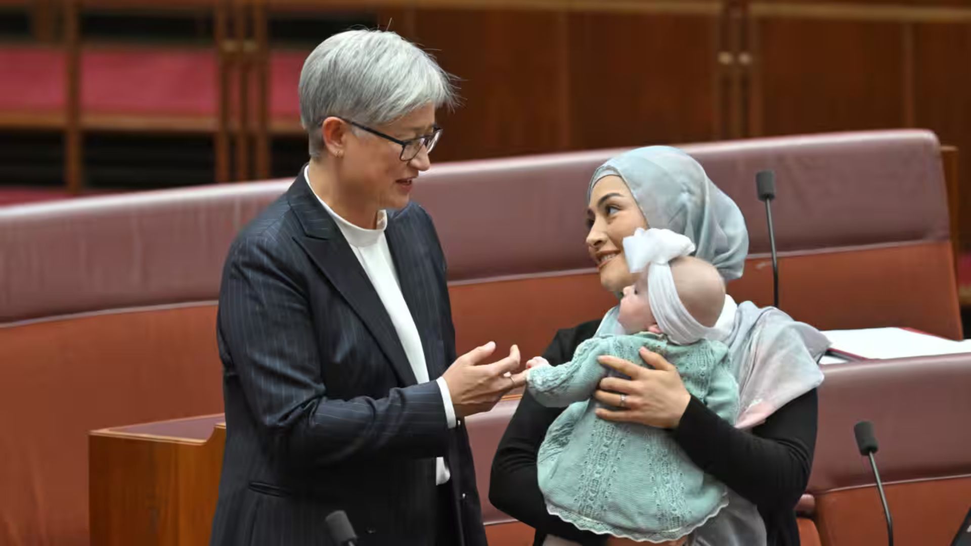 Photo of Senators Penny Wong and Fatima Payman in the Senate chamber. Payman is holding her baby and smiling.